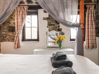 A bedroom with a white bed and gray towels on it with windows and a white dresser with a mirror and yellow flowers at Honey Bee Cottage in Bradworthy