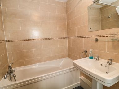 A bathroom with a bathtub a sink with two taps a wall-mounted mirror and beige tiled walls at Honey Bee Cottage in Bradworthy