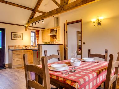 A dining area with a table set for two near a kitchen with wooden cabinets and exposed wooden beams at Woodpecker Cottage in Bradworthy near Bude