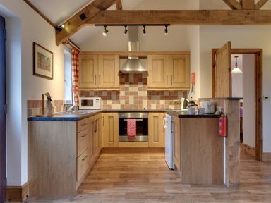 A kitchen with wooden cabinets tiled backsplash an oven a microwave a sink and a fire extinguisher at Woodpecker Cottage in Bradworthy near Bude