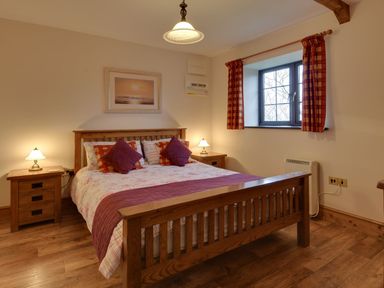 A bedroom with a wooden bed frame two bedside tables two lamps a window with red curtains and a framed picture above the bed at Woodpecker Cottage in Bradworthy near Bude
