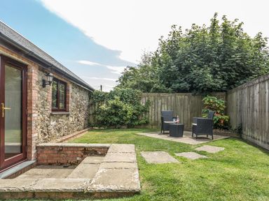 A small garden with stone steps leading to a door a brick and stone wall a patio with two chairs and a table with drinks and wooden fencing at Woodpecker Cottage in Bradworthy near Bude