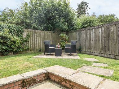 A garden with two black chairs and a table with wine glasses and a bottle surrounded by a wooden fence and greenery at Woodpecker Cottage in Bradworthy near Bude