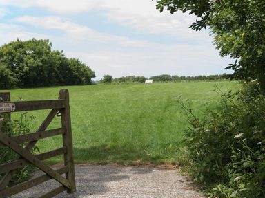 A green field with a wooden gate in the foreground and a white bench in the distance at Woodpecker Cottage in Bradworthy near Bude