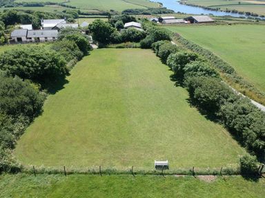 A large fenced grassy field with a white bench near the fence surrounded by trees and buildings in the background at Wagtail Bradworthy near Bude