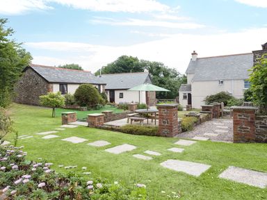 A garden with paved path and outdoor seating under umbrella near stone and brick cottages at Wagtail Bradworthy near Bude