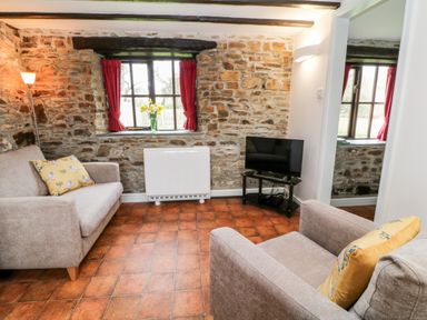 A living room with stone walls a window with red curtains two sofas with yellow pillows a television on a stand and a floor lamp at Wagtail in Bradworthy near Bude