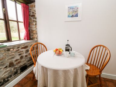 A dining area with a round table covered with a cloth two wooden chairs a bowl of fruit a coffee press and two mugs on the table a window with red curtains and a stone wall at Wagtail in Bradworthy near Bude