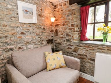 A seating area with a beige sofa and a yellow cushion near a window with red curtains and flowers on the windowsill at Wagtail Bradworthy near Bude