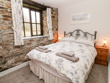 A bedroom with a double bed between two wooden bedside tables with lamps and a window with floral curtains at Wagtail in Bradworthy near Bude