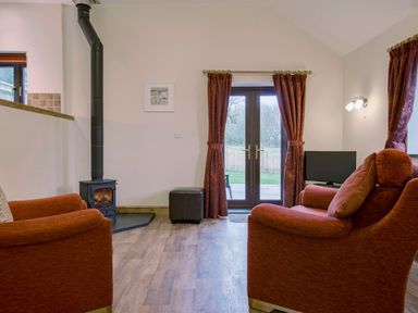 A living room with two armchairs a wood burning stove and glass doors with curtains at Jenny Wren in Bradworthy