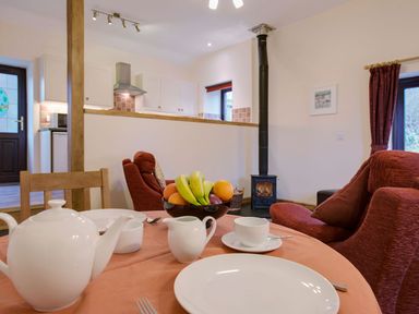 A dining table with white teapot cups and a bowl of fruit near red armchairs and a wood stove in a kitchen and living room at Jenny Wren in Bradworthy