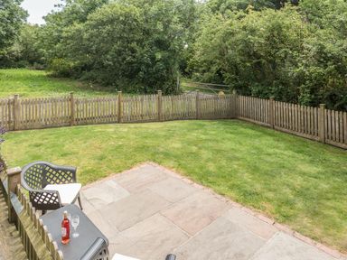 A fenced grassy garden with a patio area and two chairs with a table and bottle at Jenny Wren in Bradworthy