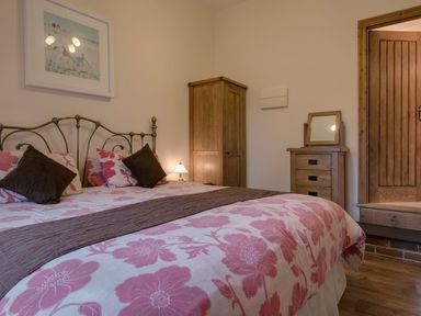 A bedroom with a double bed with floral bedding two brown pillows a wooden wardrobe a chest with mirror and a wooden door at Jenny Wren in Bradworthy