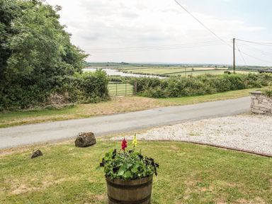 A garden with a flower planter and rocks next to a road with fields and a lake in the background at Jenny Wren in Bradworthy