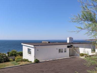 A flat roof white house overlooking the sea with a driveway and some greenery at Boscregan in Porthleven