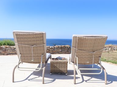 Two wicker lounge chairs and a wicker side table on a stone patio overlooking the ocean at Boscregan in Porthleven