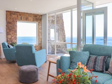 A living room with blue armchairs a sofa flowers a stone wall and ocean view at Boscregan in Porthleven