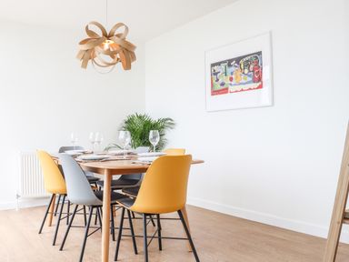 A dining room with a wooden table set with plates and glasses surrounded by yellow and gray chairs a modern wooden light fixture and a framed picture on the wall at Boscregan in Porthleven
