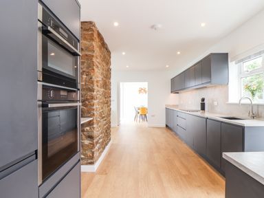 A kitchen with dark cabinets a stone wall wooden floor and a window at Boscregan in Porthleven