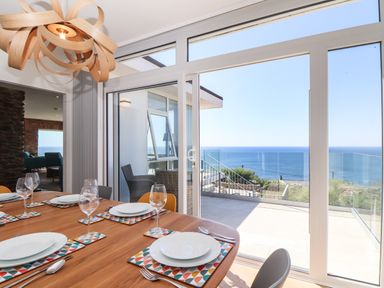 A dining room with a wooden table set with plates and wine glasses next to large glass doors overlooking a sea view at Boscregan in Porthleven