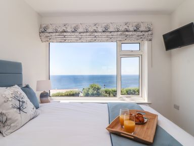 A bedroom with a bed facing a large window showing a sea view a wooden tray with glasses of orange juice and a croissant on the bed a lamp on a bedside table and a wall-mounted television