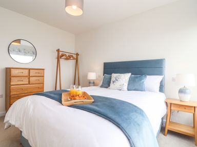 A bedroom with a bed and blue headboard a wooden dresser a wooden clothes rack with hangers and bedside tables with lamps at Boscregan in Porthleven