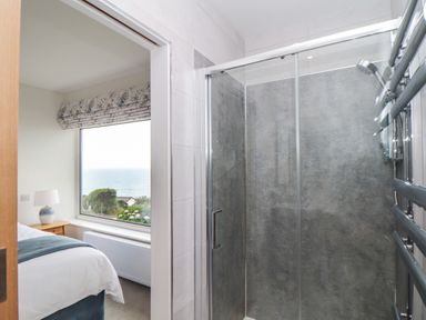A bathroom with a glass shower enclosure next to a bedroom with a large window overlooking the sea at Boscregan in Porthleven