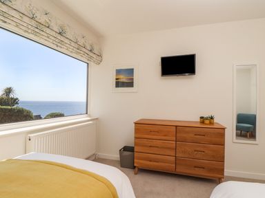 A bedroom with a wooden dresser a wall-mounted TV a window showing sea view and a mirror at Boscregan in Porthleven