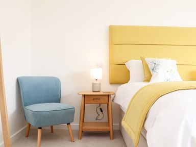 A bedroom with a blue chair beside a wooden nightstand with a lamp next to a bed with a yellow headboard and bedding at Boscregan in Porthleven