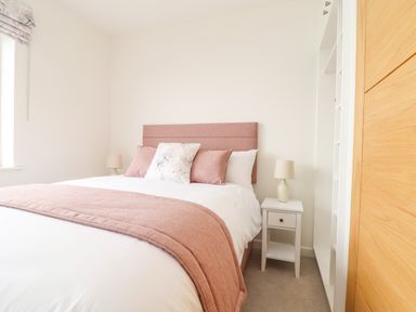 A bedroom with a bed a pink headboard pink and white pillows a pink throw and a white bedside table with a lamp at Boscregan in Porthleven