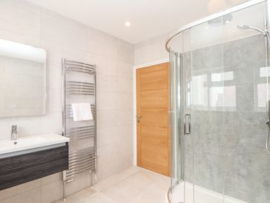 A bathroom with a wooden door a wall-mounted sink a mirror a towel radiator and a curved glass shower enclosure at Boscregan in Porthleven