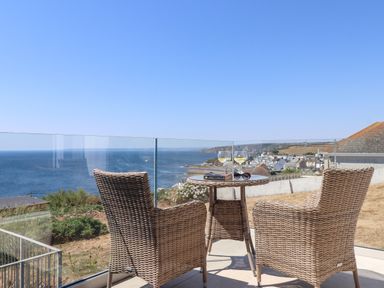 A balcony with two wicker chairs and a round table overlooking the ocean and a coastal town at Boscregan in Porthleven