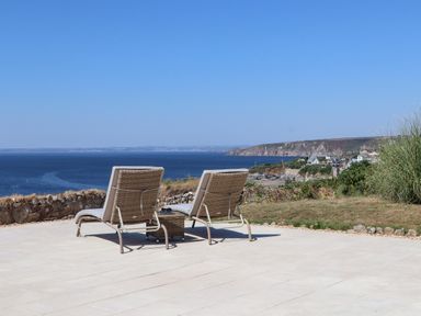 Two lounge chairs on a tiled patio facing the sea with a coastline and houses in the background at Boscregan in Porthleven