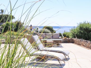 An outdoor patio with lounge chairs a sofa and a table overlooking the sea with plants and a stone wall surrounding the area at Boscregan in Porthleven