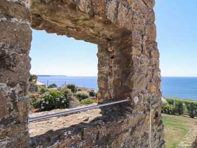 A stone wall with a rectangular opening showing a garden with flowers houses and a sea view at Boscregan Porthleven