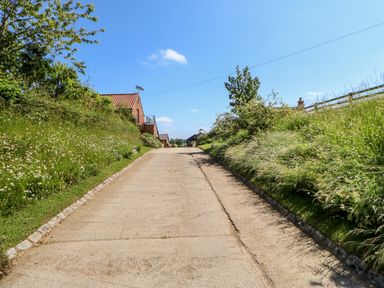 A paved driveway with grassy banks and houses on the left side at Lovesome Pod Lovesome Hill near Northallerton
