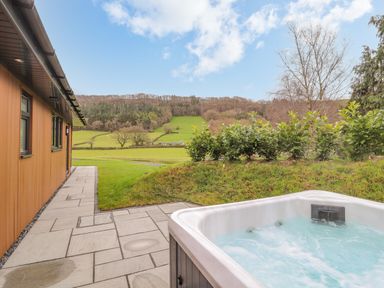 An outdoor hot tub on a stone patio next to a wooden building overlooking grassy hills and trees at Pool View in Meifod Powys
