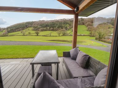 A covered outdoor patio with a glass table and wicker seating cushions overlooking fields and hills at Pool View in Meifod Powys