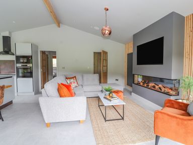 A living room with a grey sectional sofa with orange and floral cushions a rectangular coffee table and an orange armchair next to a modern fireplace and mounted TV at Pool View in Meifod Powys