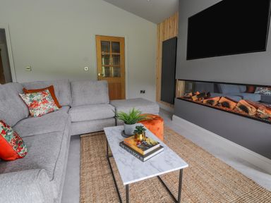 A living room with a gray sectional sofa with floral and orange cushions a marble coffee table with books and plants a wall-mounted TV and an electric fireplace at Pool View in Meifod Powys
