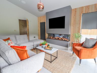 A living room with a grey sectional sofa with orange and floral cushions an orange armchair a low rectangular coffee table a wall-mounted tv and a modern fireplace at Pool View in Meifod Powys