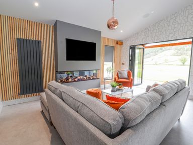 A living room with a gray sectional sofa orange chairs a wall-mounted TV and bi-fold doors opening to a garden at Pool View in Meifod Powys