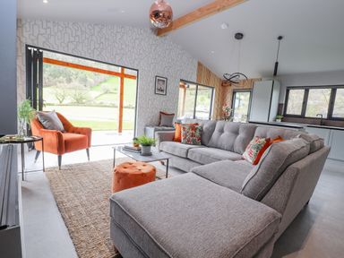 A living room with a gray sectional sofa floral and orange pillows an orange armchair a round orange ottoman a coffee table and large windows showing a green landscape at Pool View in Meifod Powys