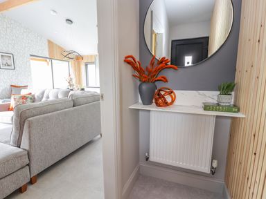 A small corner with a white shelf above a radiator holding a vase with orange dried flowers a decorative orange orb a small green plant and a book with a round mirror above at Pool View in Meifod Powys