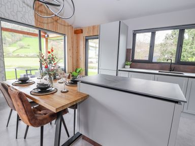 A kitchen area with a wooden dining table set with black plates and glassware next to a kitchen island and windows showing an outdoor view at Pool View in Meifod Powys