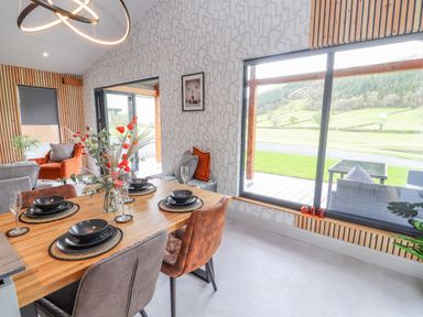 A dining area with a wooden table set for four people and large windows showing a green landscape at Pool View in Meifod Powys