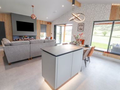 A living room with a grey sofa a wall mounted TV a dining table with chairs and flowers on it and large windows at Pool View in Meifod Powys