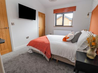 A bedroom with a bed covered with white and orange bedding a black nightstand with a vase and flowers a window and a wall mounted tv at Pool View Meifod Powys