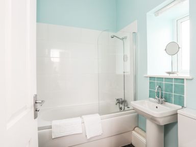 A bathroom with a bathtub shower combination a pedestal sink a mirror on the window sill and a towel on the bathtub at Plas Newydd in Llanfaelog near Rhosneigr